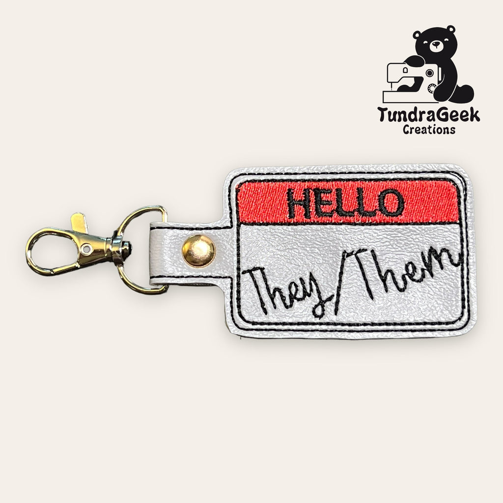 A keychain featuring a stitched Hello name tag design with They/Them written in black script, next to the TundraGeek Creations logo with a bear and sewing machine.
