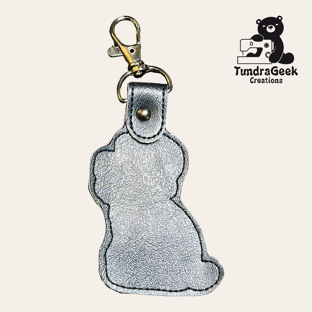 Silver metallic keychain shaped like a sitting animal, possibly a dog or cat, with a clasp. TundraGeek Creations logo featuring a bear and sewing machine in the top right corner.