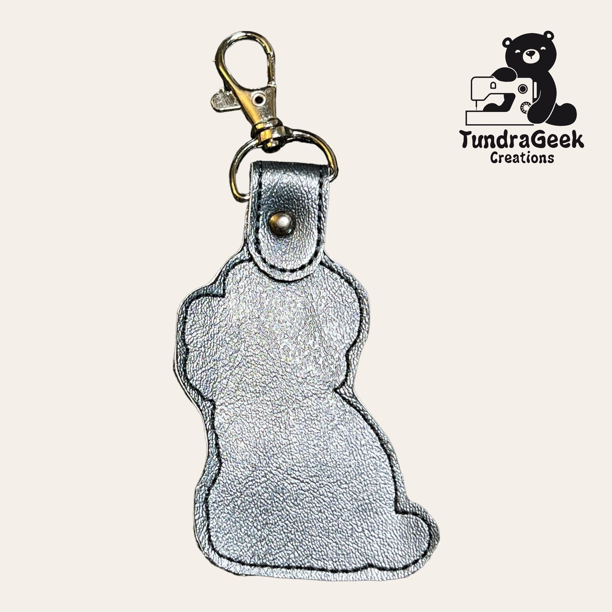 Silver metallic keychain shaped like a sitting animal, possibly a dog or cat, with a clasp. TundraGeek Creations logo featuring a bear and sewing machine in the top right corner.