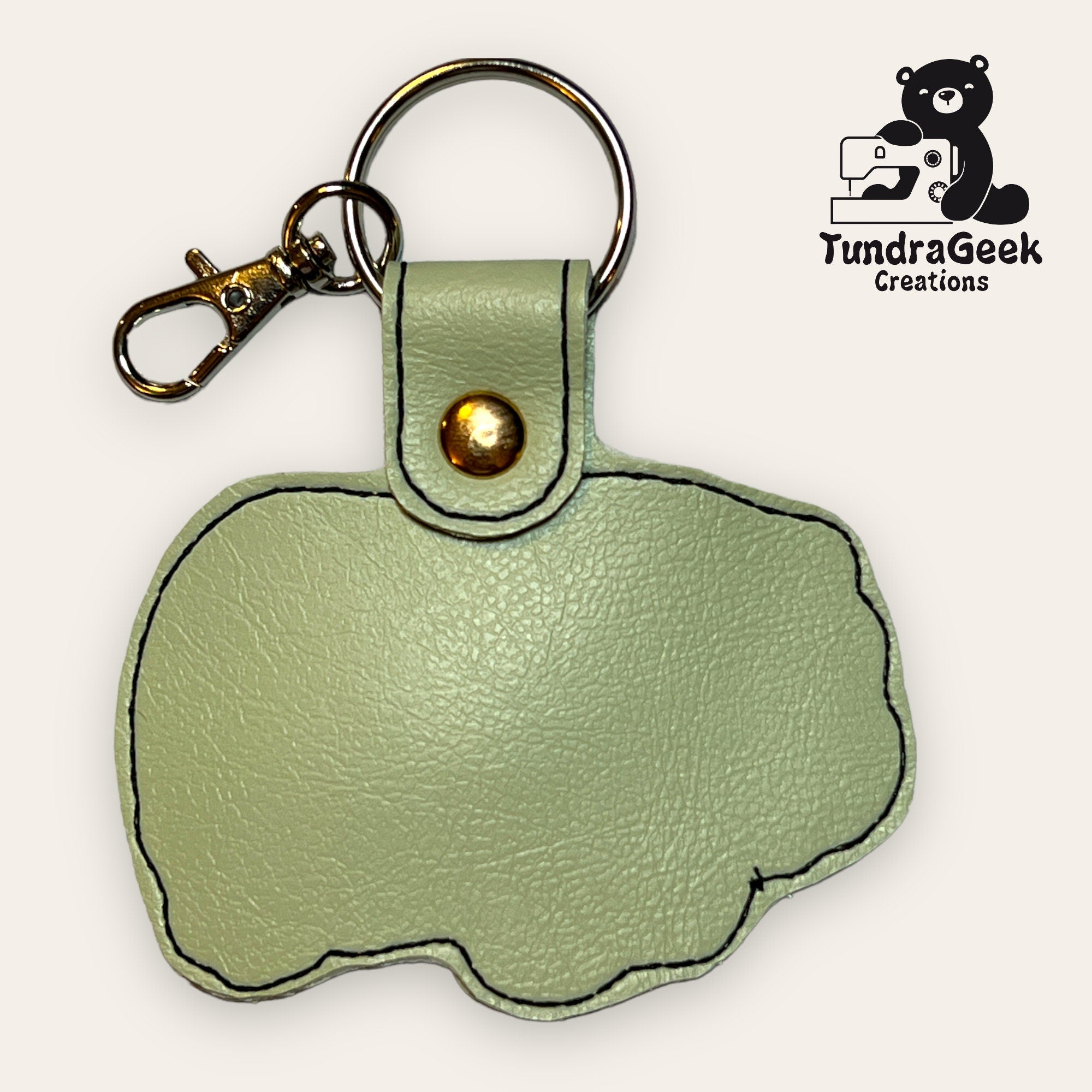 Light green faux leather keychain shaped like a hippopotamus, with a gold snap and metal keyring. TundraGeek Creations logo with a bear and sewing machine is in the top right corner.