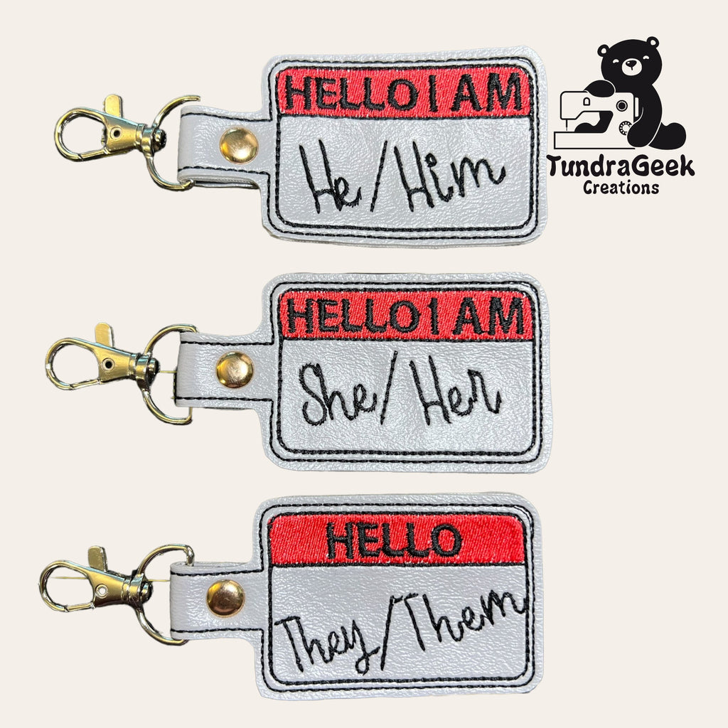 Three embroidered keychains resembling name tags display pronouns: He/Him, She/Her, and They/Them. TundraGeek Creations logo with a bear and sewing machine appears on the right.