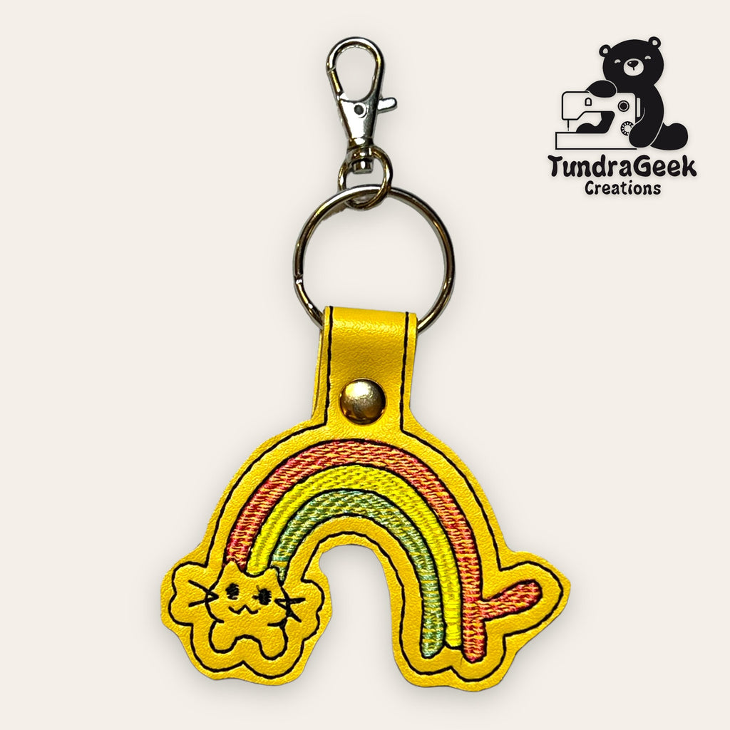Yellow keychain with an embroidered rainbow and cat face, attached to a metal ring and clip. TundraGeek Creations logo with a bear and sewing machine is in the top right corner.