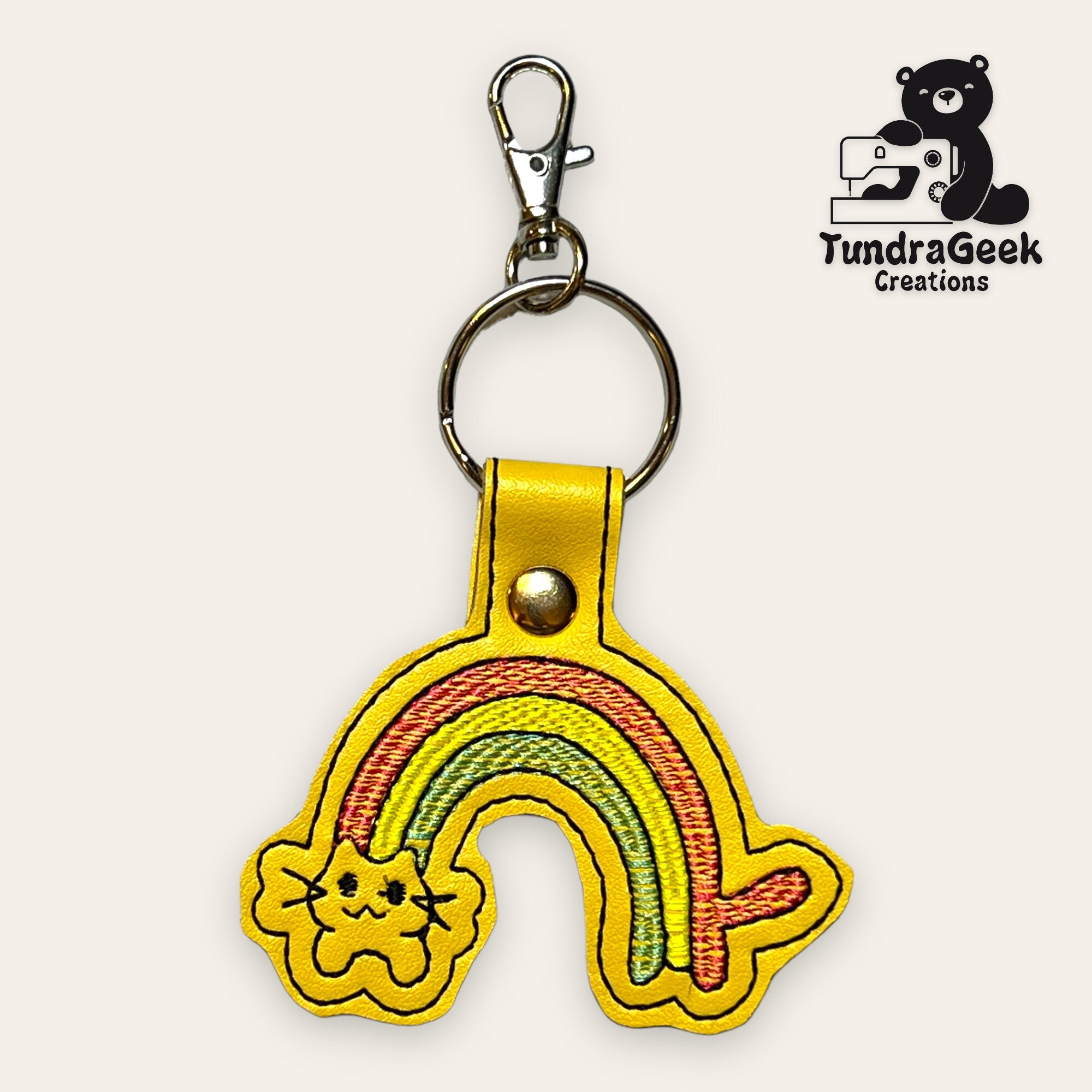 Yellow keychain with an embroidered rainbow and cat face, attached to a metal ring and clip. TundraGeek Creations logo with a bear and sewing machine is in the top right corner.