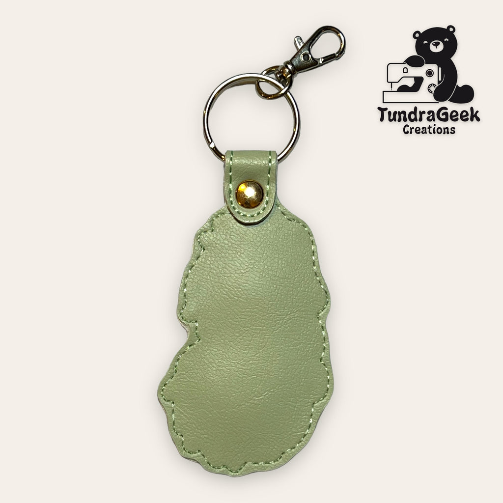 Light green faux leather keychain with irregular edges, featuring a gold rivet and metal clasp. TundraGeek Creations logo with a bear and sewing machine is in the top right corner.