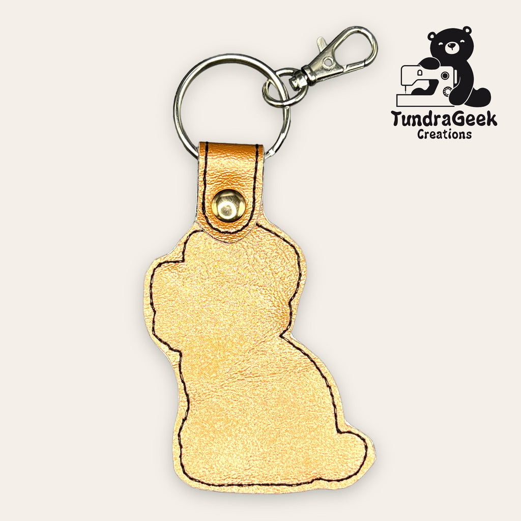 A gold leather keychain shaped like a sitting cat, with a metal clasp and keyring. TundraGeek Creations logo featuring a bear and sewing machine is in the top right corner.