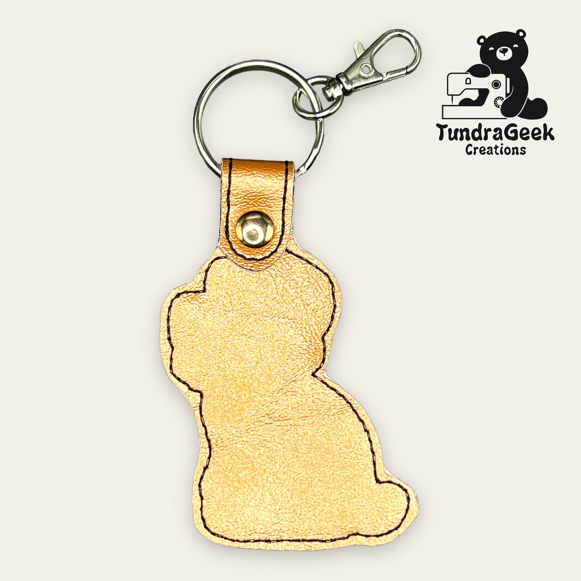 A gold leather keychain shaped like a sitting cat, with a metal clasp and keyring. TundraGeek Creations logo featuring a bear and sewing machine is in the top right corner.