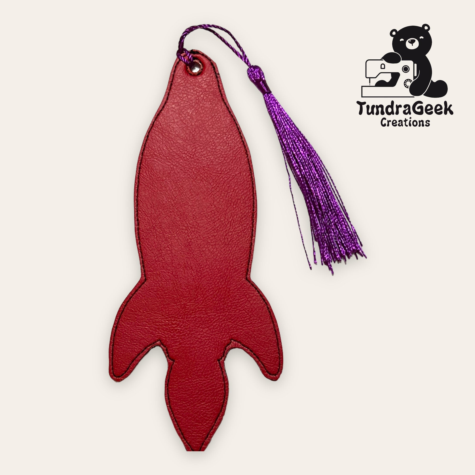 Red rocket-shaped leather bookmark with a purple tassel, displayed on a light background. TundraGeek Creations logo featuring a bear and sewing machine is in the top right corner.
