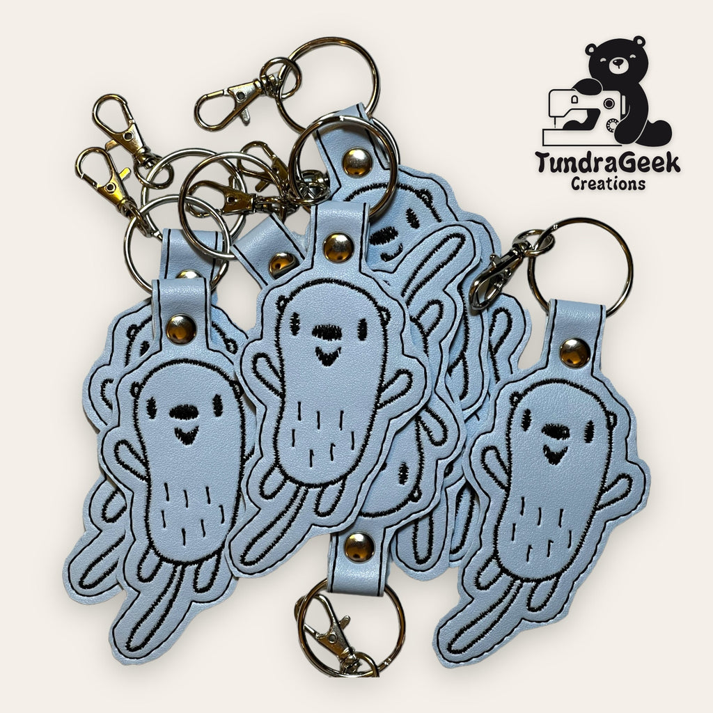 A pile of embroidered keychains shaped like smiling cartoon bacteria, with metal rings and clips. TundraGeek Creations logo is in the top right corner.
