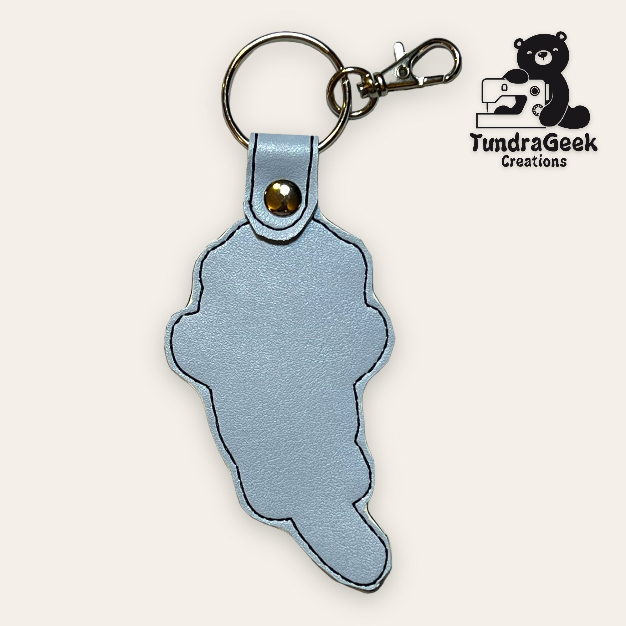 A light blue leather keychain in the shape of Michigans Upper Peninsula, attached to a metal key ring and clasp. TundraGeek Creations logo is visible in the top right corner.