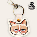 A keychain with an embroidered grumpy cat face on white faux leather, featuring a metal key ring and clasp. TundraGeek Creations logo is in the upper right corner.