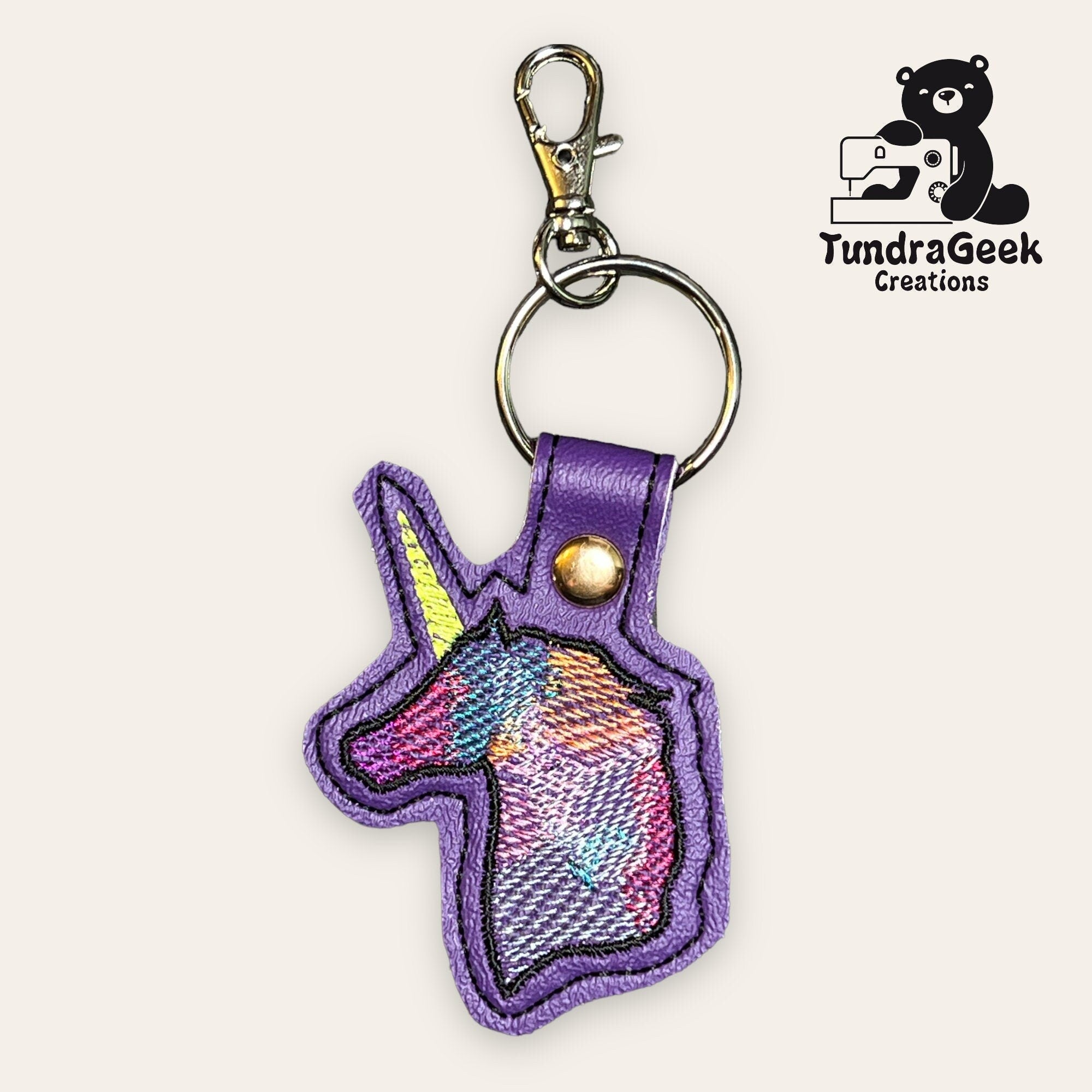 A multicolored embroidered unicorn head keychain with a purple border and metal clasp, displayed next to the TundraGeek Creations logo.
