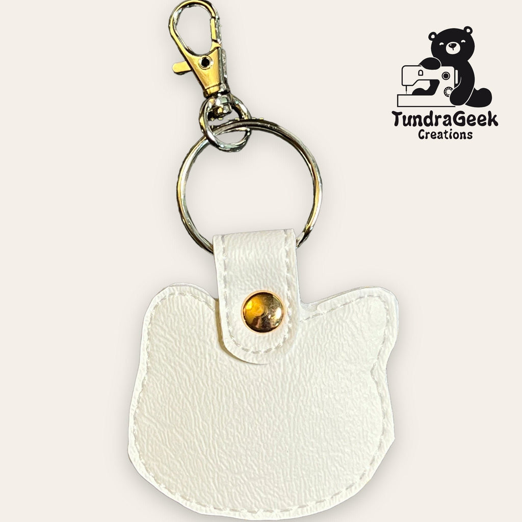 White cat-shaped faux leather keychain with gold snap and metal clip, displayed on a light background with the TundraGeek Creations logo in the corner.