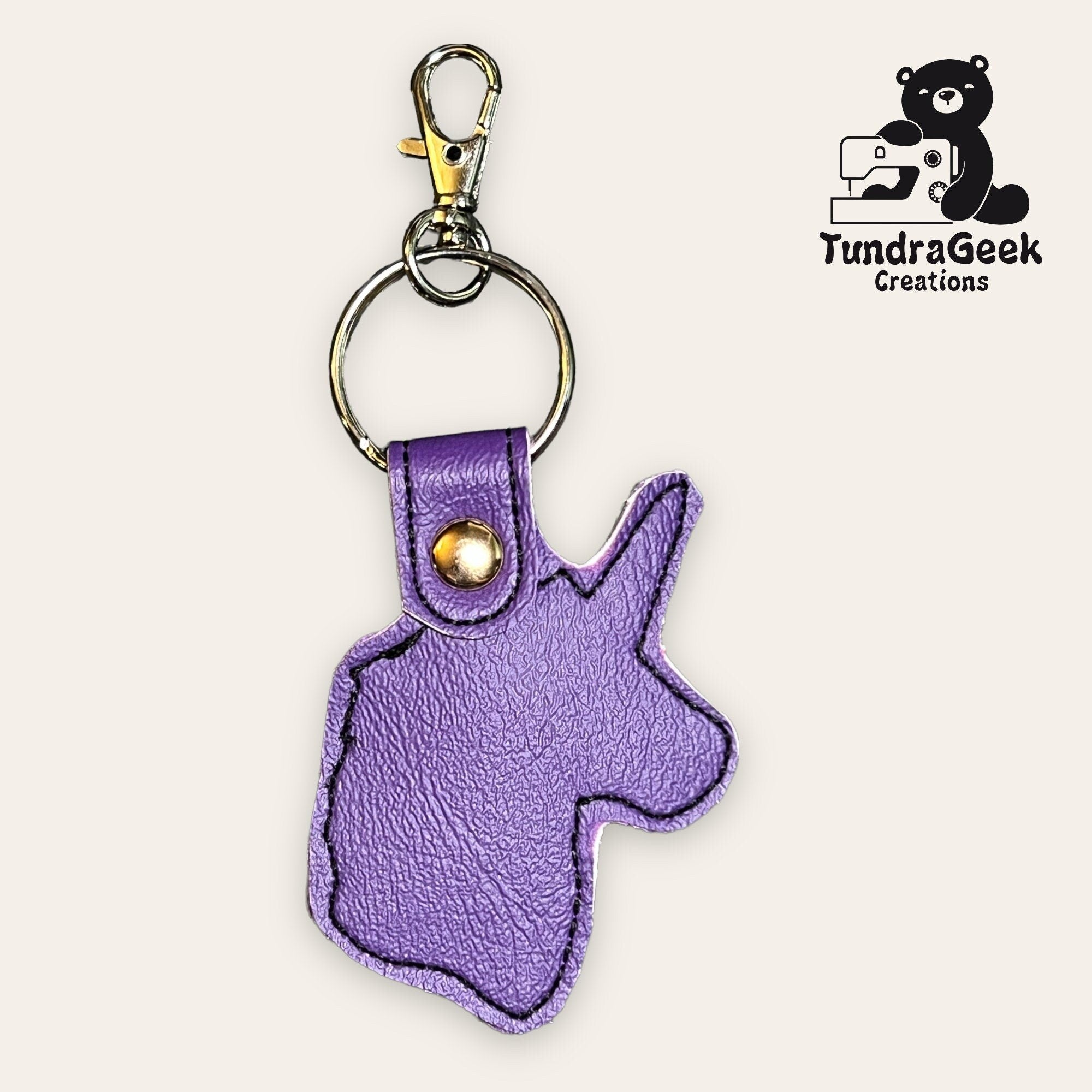 Purple faux leather keychain shaped like a unicorn head with a gold snap and metal keyring, displayed next to the TundraGeek Creations logo.