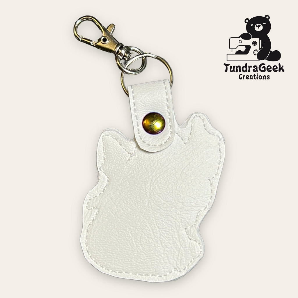 White faux leather keychain with an animal-shaped outline, metal snap, and clasp. TundraGeek Creations logo with a bear holding a sewing machine in the top right corner.