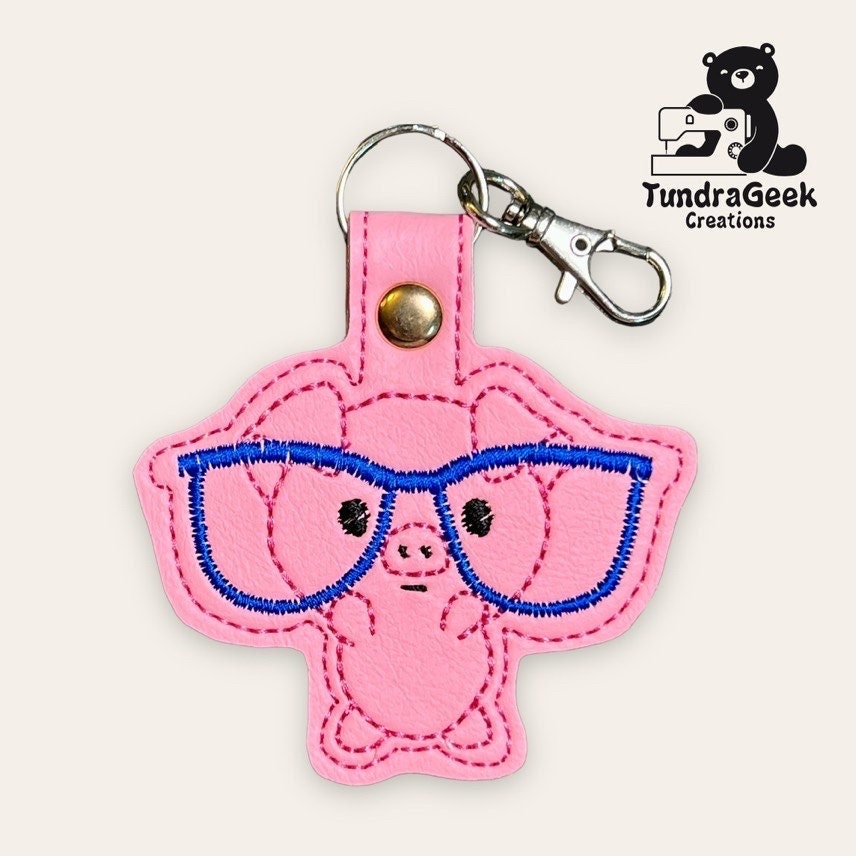 Pink pig keychain with blue embroidered glasses and a metal clasp, displayed on a light background with the TundraGeek Creations logo in the corner.