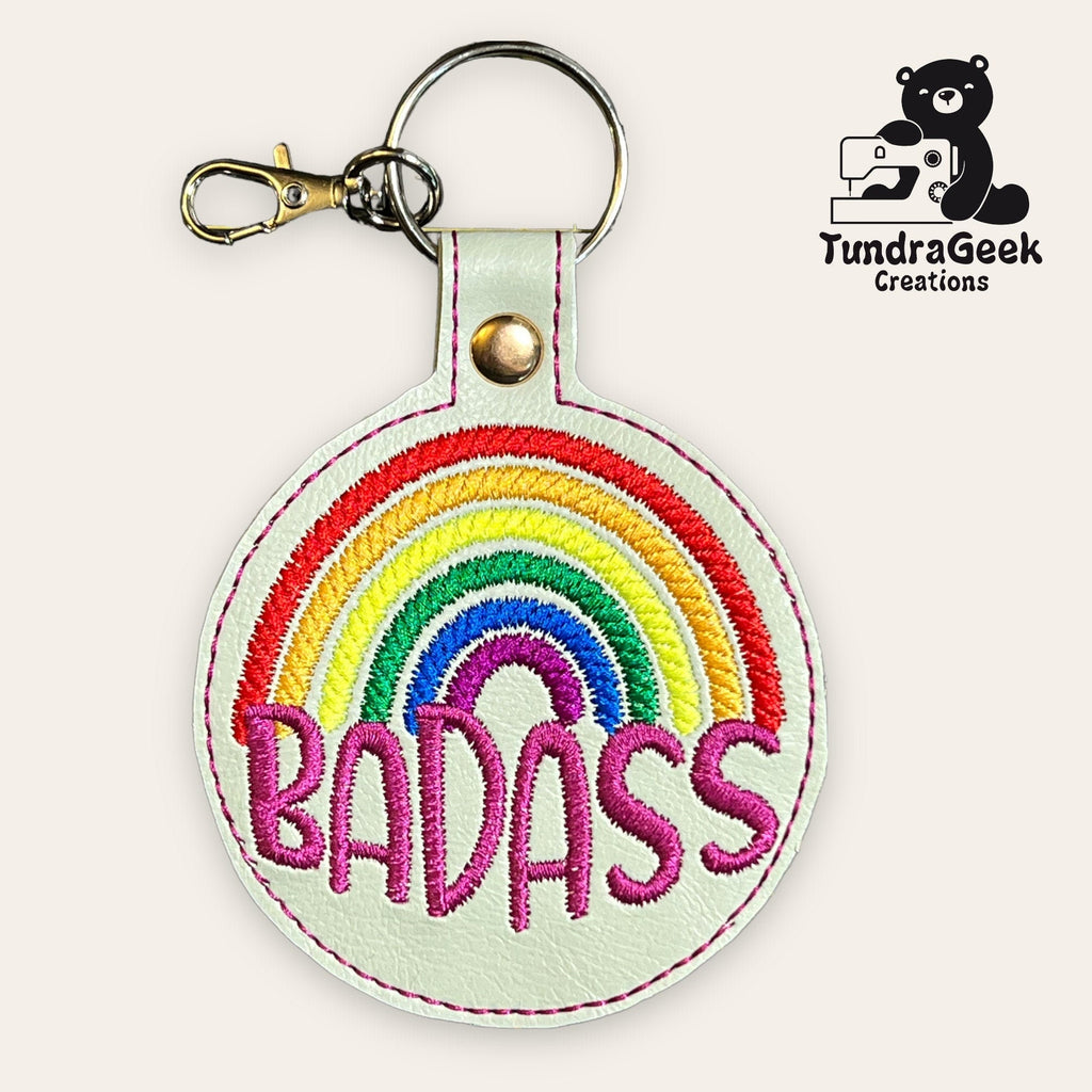 Round white keychain with an embroidered rainbow and the word BADASS in bold pink letters. TundraGeek Creations logo with bear and sewing machine is in the top right corner.