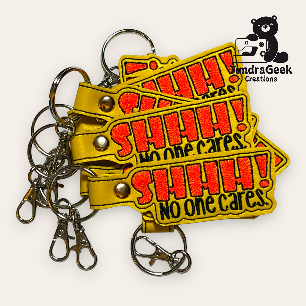Four yellow keychains with red and black embroidered text reading SHHH! No one cares. are stacked, accompanied by metal clips and labeled with the TundraGeek Creations logo.
