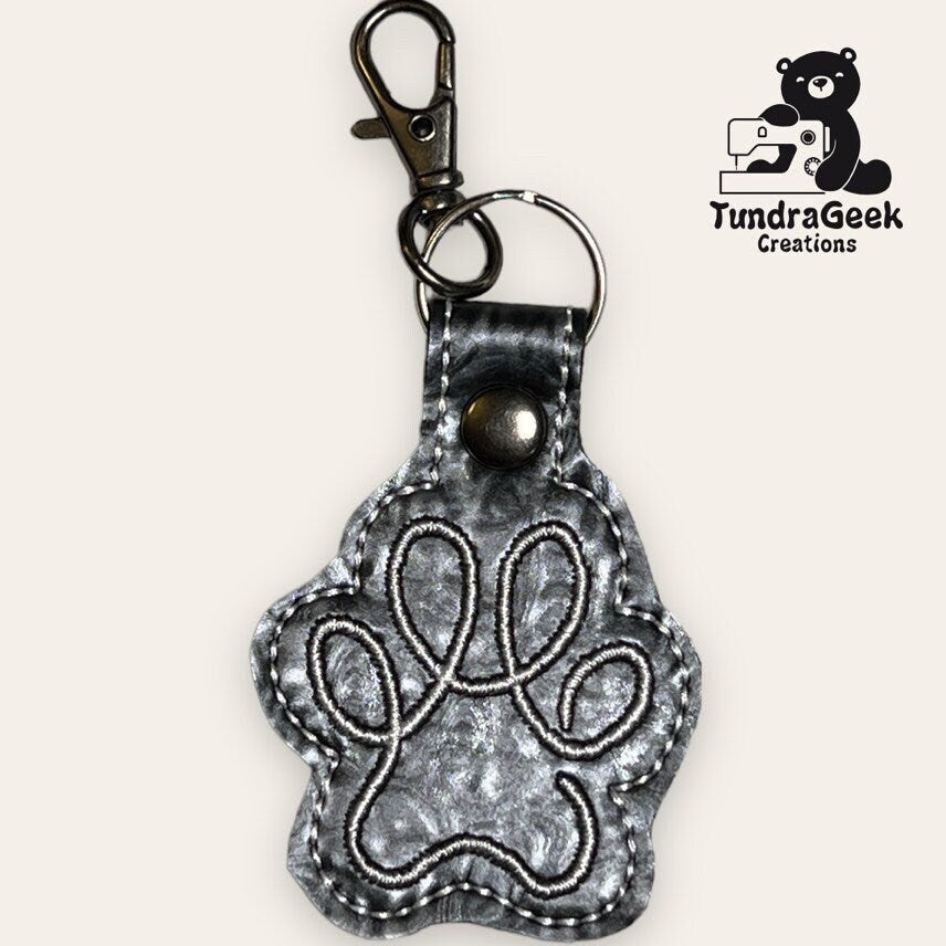 A metallic paw-shaped keychain with a stitched paw print design, metal clasp, and keyring. TundraGeek Creations logo is visible in the top right corner.
