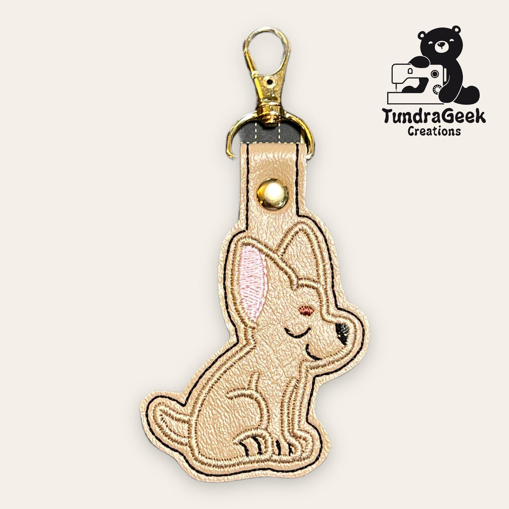 Keychain featuring an embroidered tan dog with pink inner ears, attached to a gold clasp. TundraGeek Creations logo with a bear and sewing machine is in the top right corner.