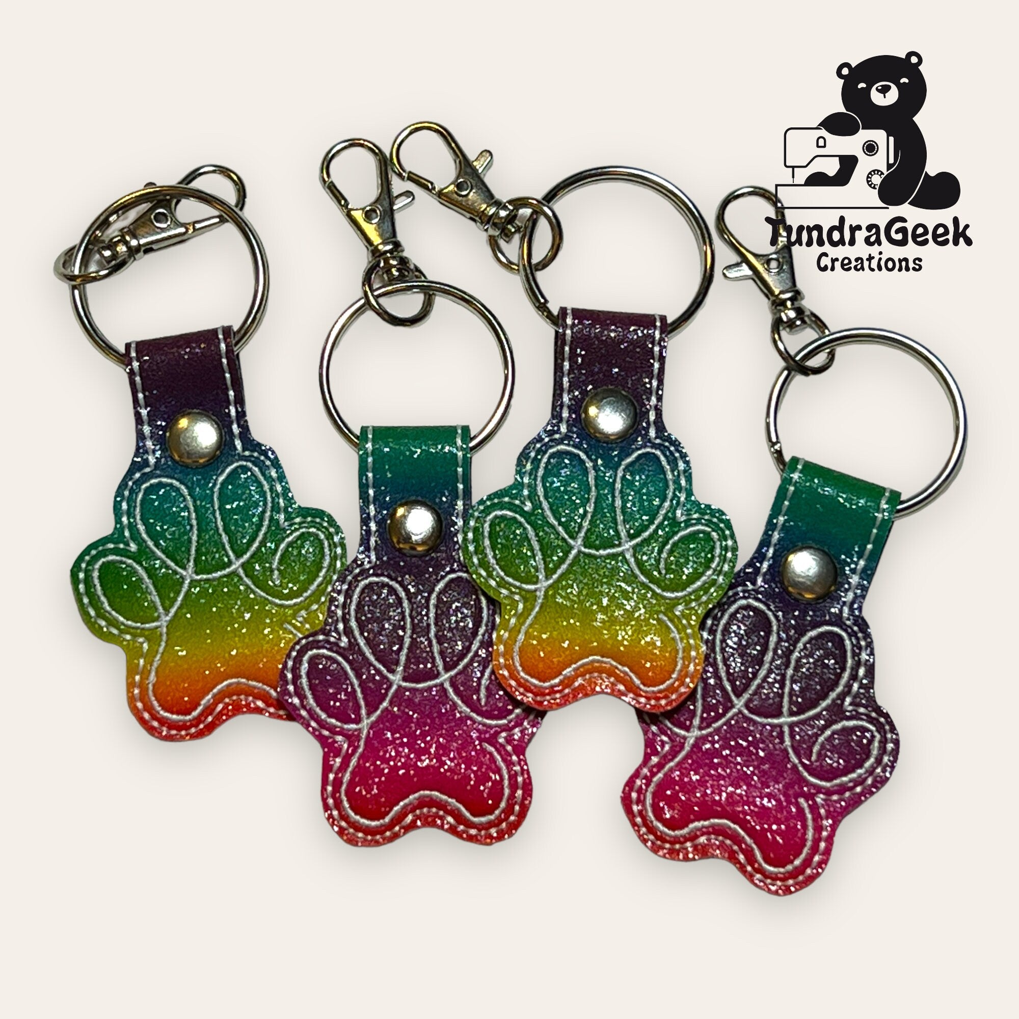 Four glittery rainbow paw print keychains with metal rings and clasps are arranged in a row. The JundraGeek Creations logo appears in the top right corner.