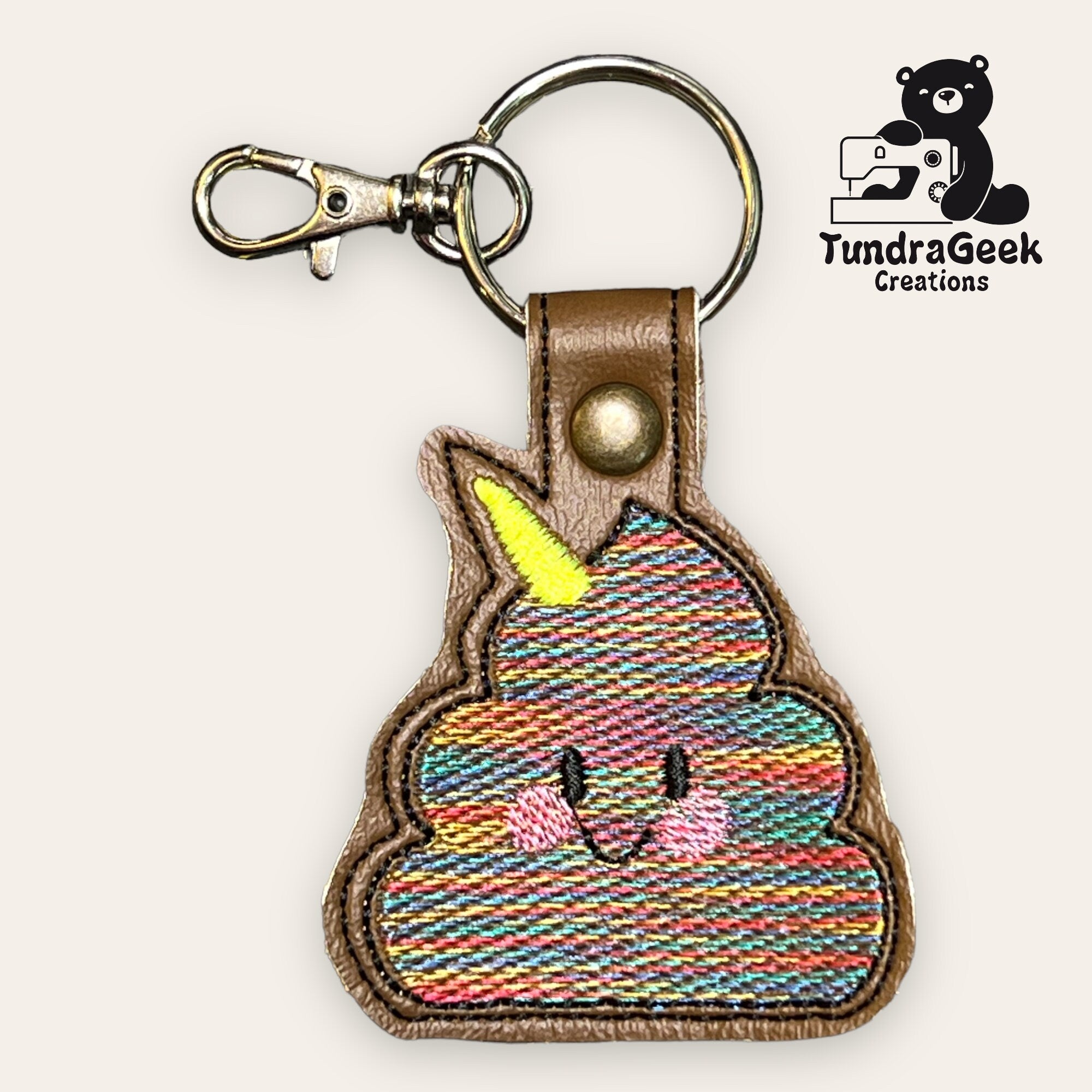 A keychain shaped like a smiling poop emoji with a yellow unicorn horn, made of colorful embroidered fabric and brown faux leather, next to the TundraGeek Creations logo.