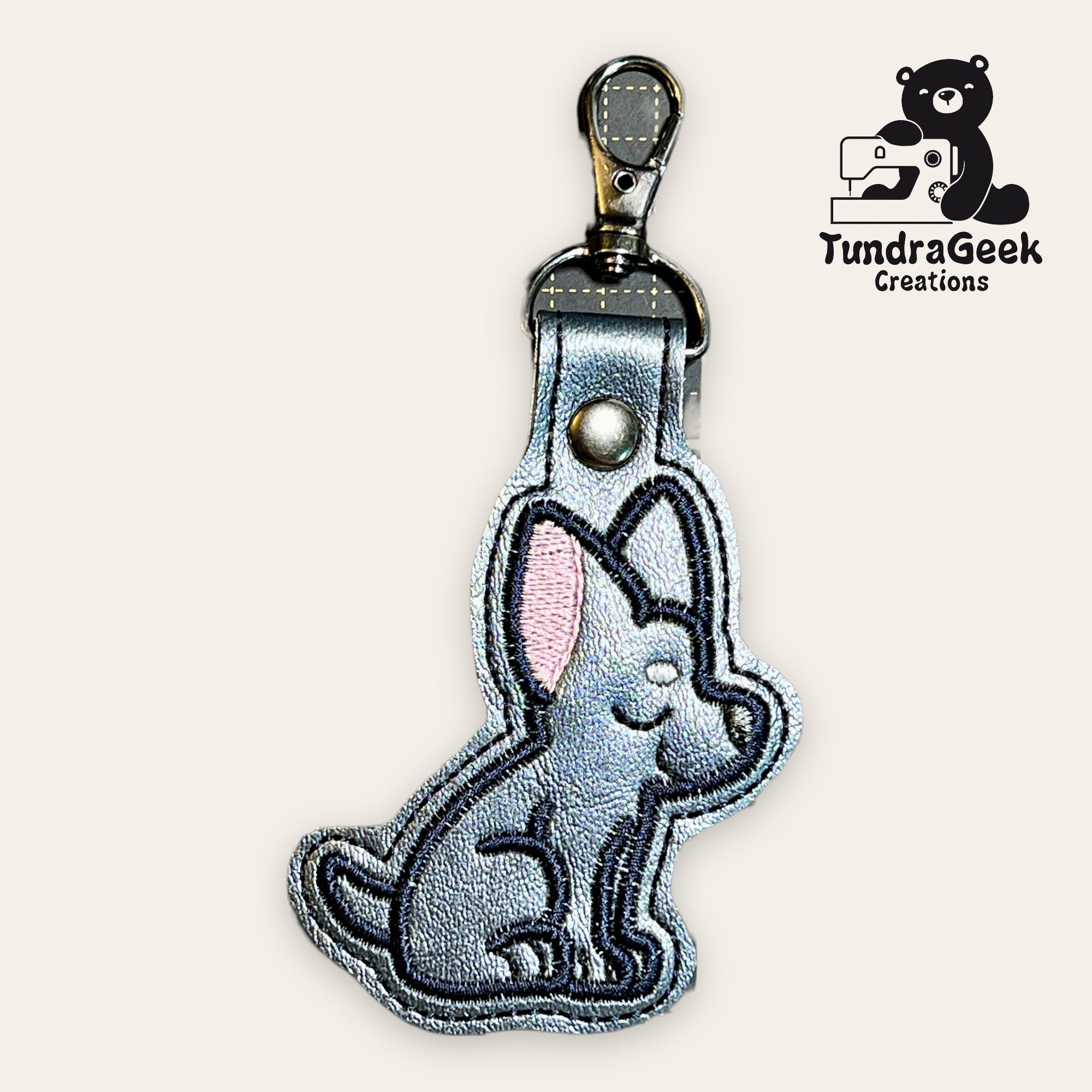 Keychain featuring a stitched cartoon dog design with a metal clasp; TundraGeek Creations logo appears in the top right corner.