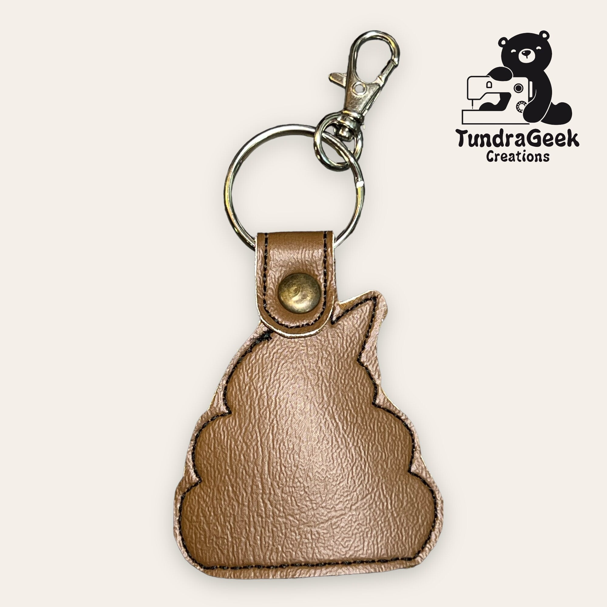 Brown faux leather keychain shaped like a poop emoji, with a metal keyring and clasp. TundraGeek Creations logo featuring a bear and sewing machine in the corner.