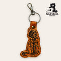Brown geometric cat-shaped keychain with black stitching and a metal clasp. TundraGeek Creations logo with a bear and sewing machine is in the top right corner.