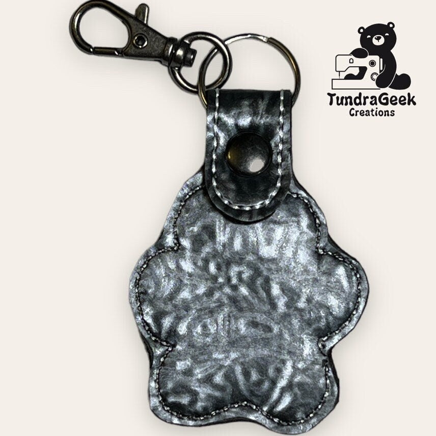 A metallic silver paw-shaped keychain with a snap button and clip, displayed next to the TundraGeek Creations logo featuring a bear with a sewing machine.