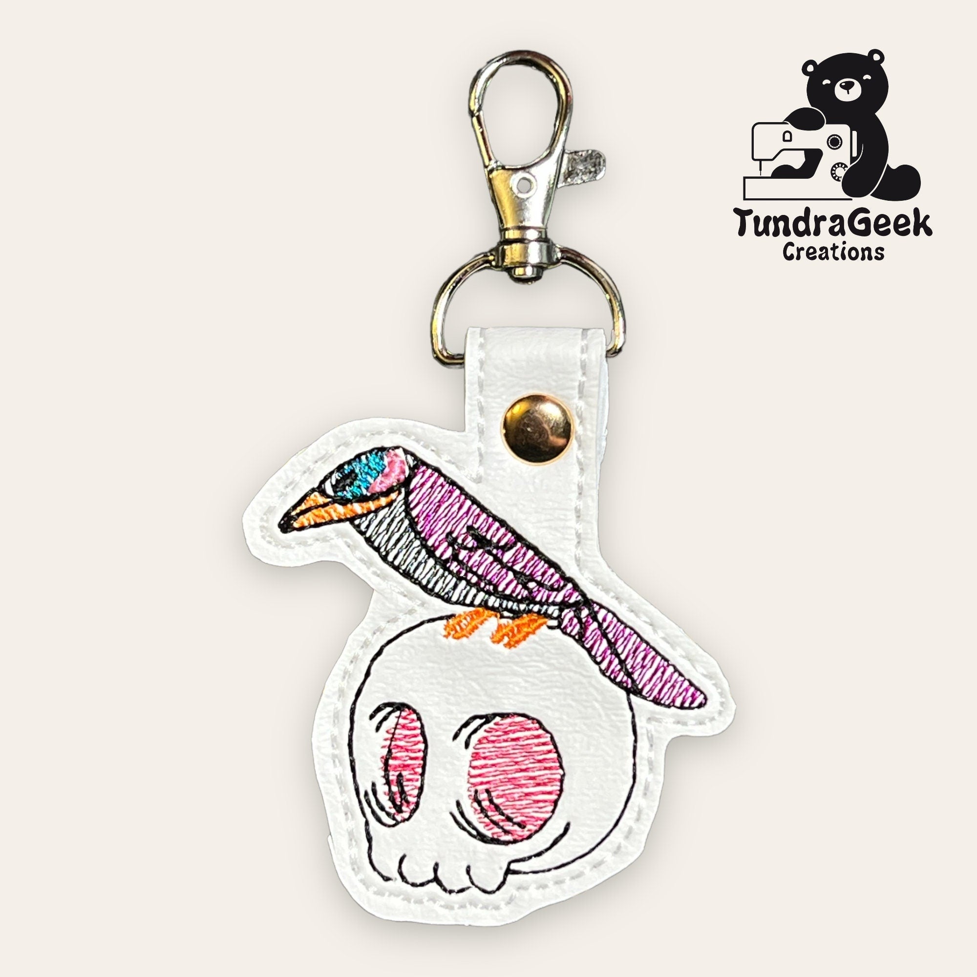 Embroidered keychain with a bird perched on a skull, attached to a metal clasp, with the TundraGeek Creations logo in the corner.