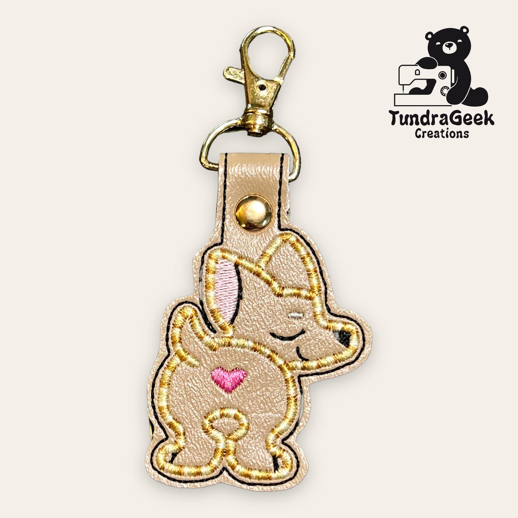 Embroidered keychain featuring a cartoon dog with a pink heart on its side and a metal clasp. TundraGeek Creations logo in the top right corner.