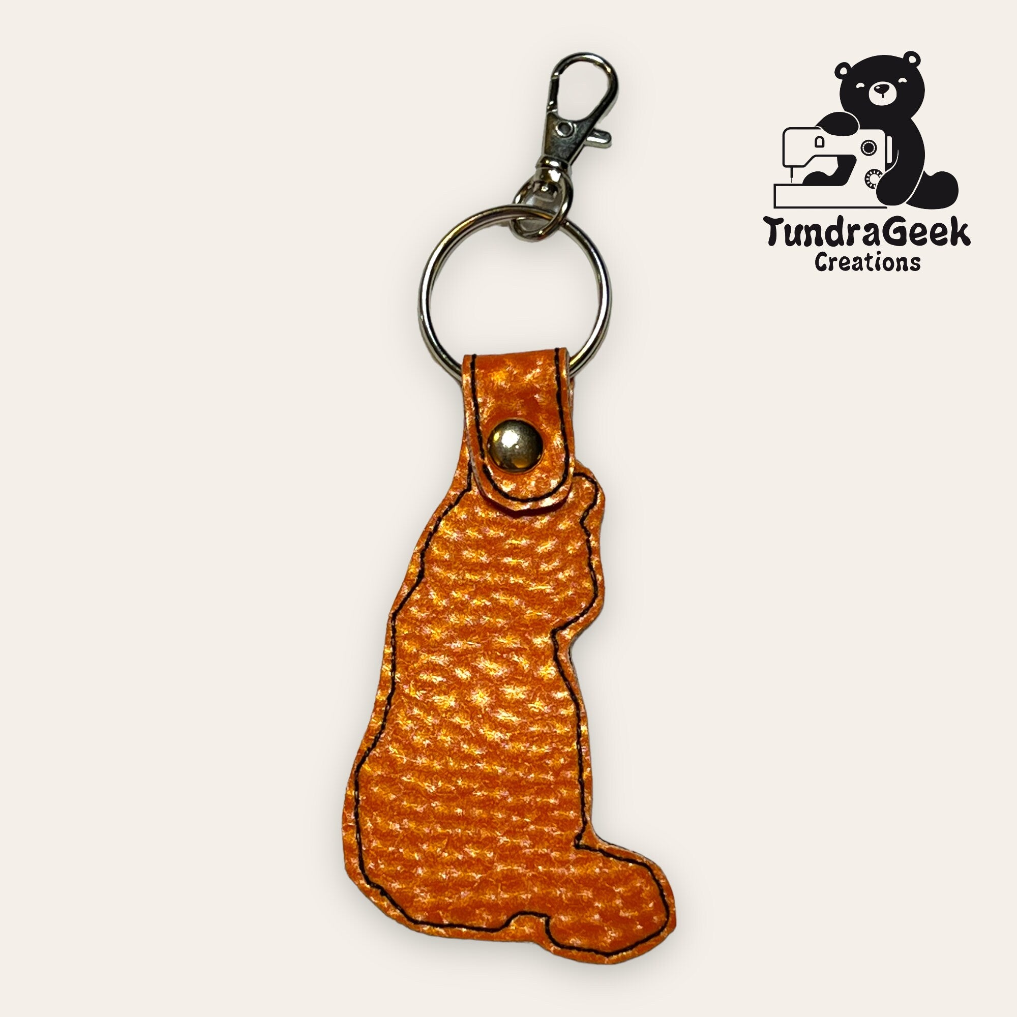 Orange, textured keychain in the shape of a sitting cat, with a metal key ring and clasp; TundraGeek Creations logo in the corner.