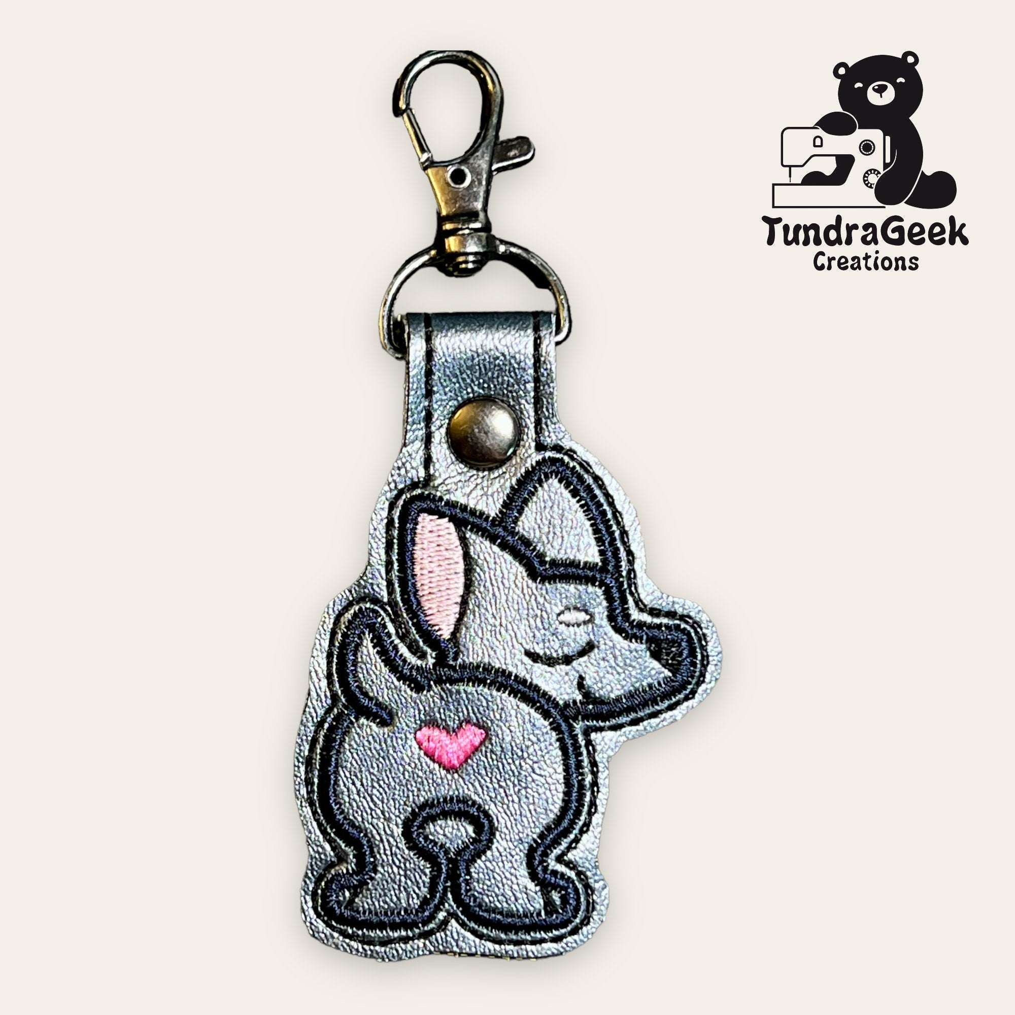 A silver keychain features an embroidered cartoon dog with a pink heart on its side, attached to a metal keyring and clasp. TundraGeek Creations logo appears in the top right corner.