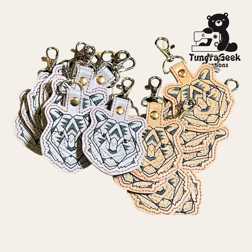 A group of embroidered tiger head keychains with metal clasps, arranged in a fan shape. TundraGeek Creations logo in the top right corner.