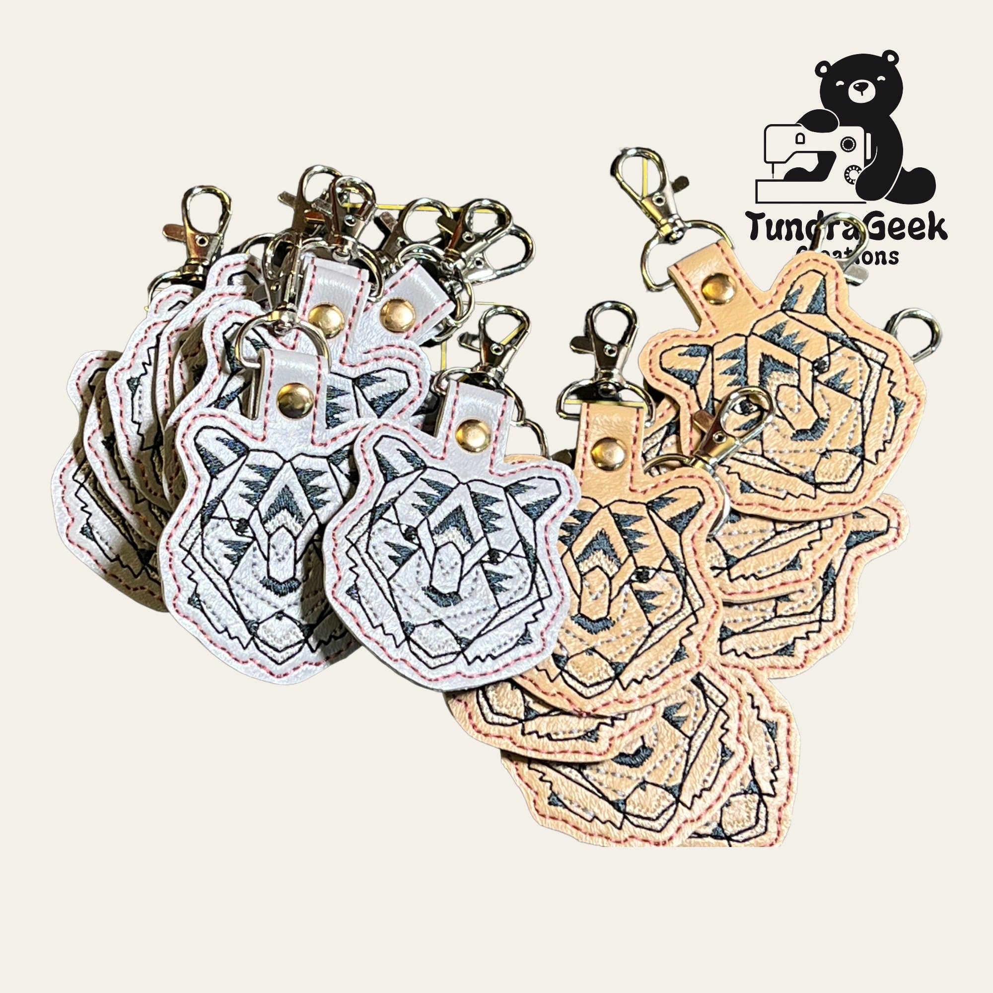 A group of embroidered tiger head keychains with metal clasps, arranged in a fan shape. TundraGeek Creations logo in the top right corner.