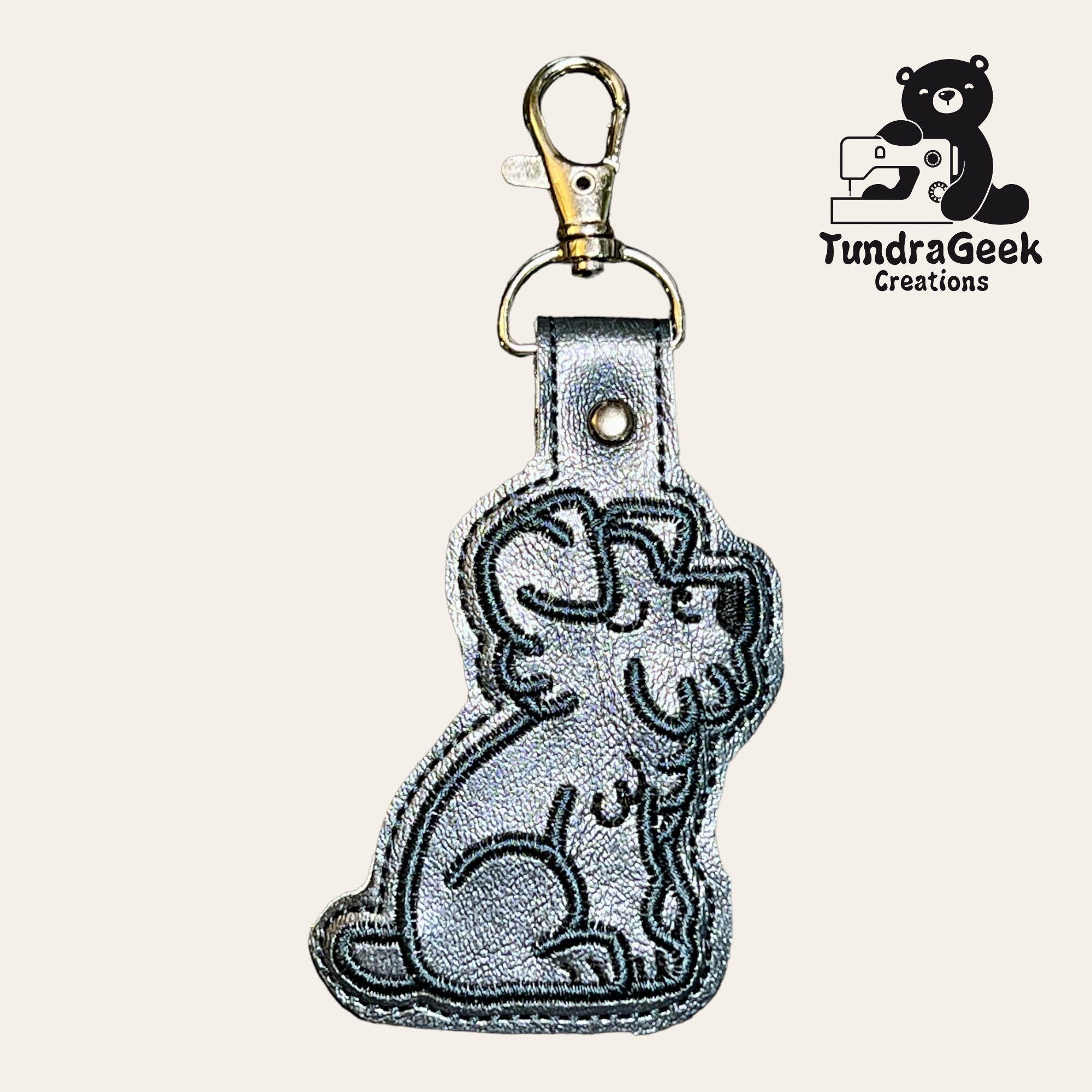 Silver dog-shaped keychain with black embroidered outline, metal clasp, and TundraGeek Creations logo featuring a bear with a sewing machine in the upper right corner.