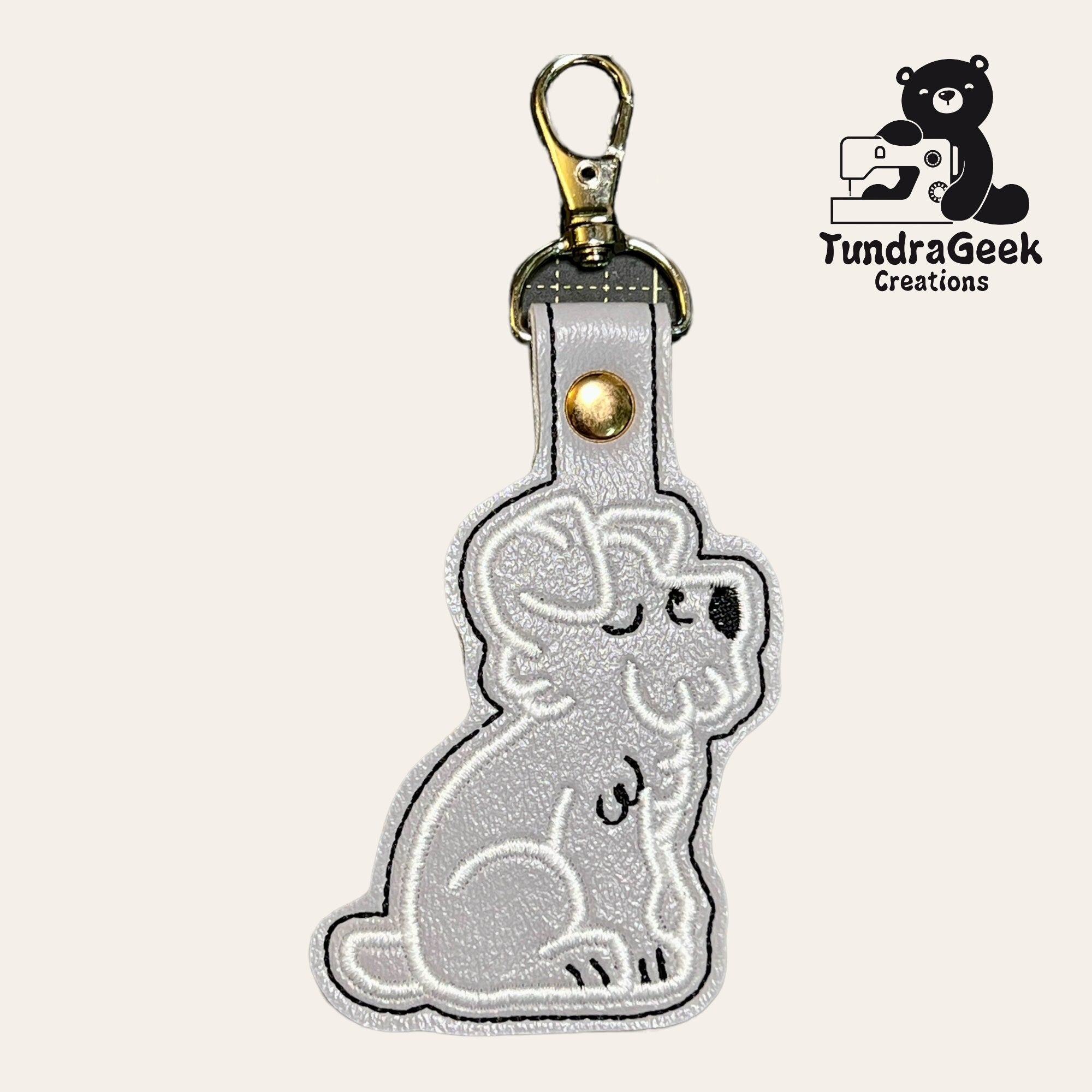 A gray embroidered keychain in the shape of a sitting koala, with a metal clasp. TundraGeek Creations logo featuring a bear and sewing machine is in the upper right corner.