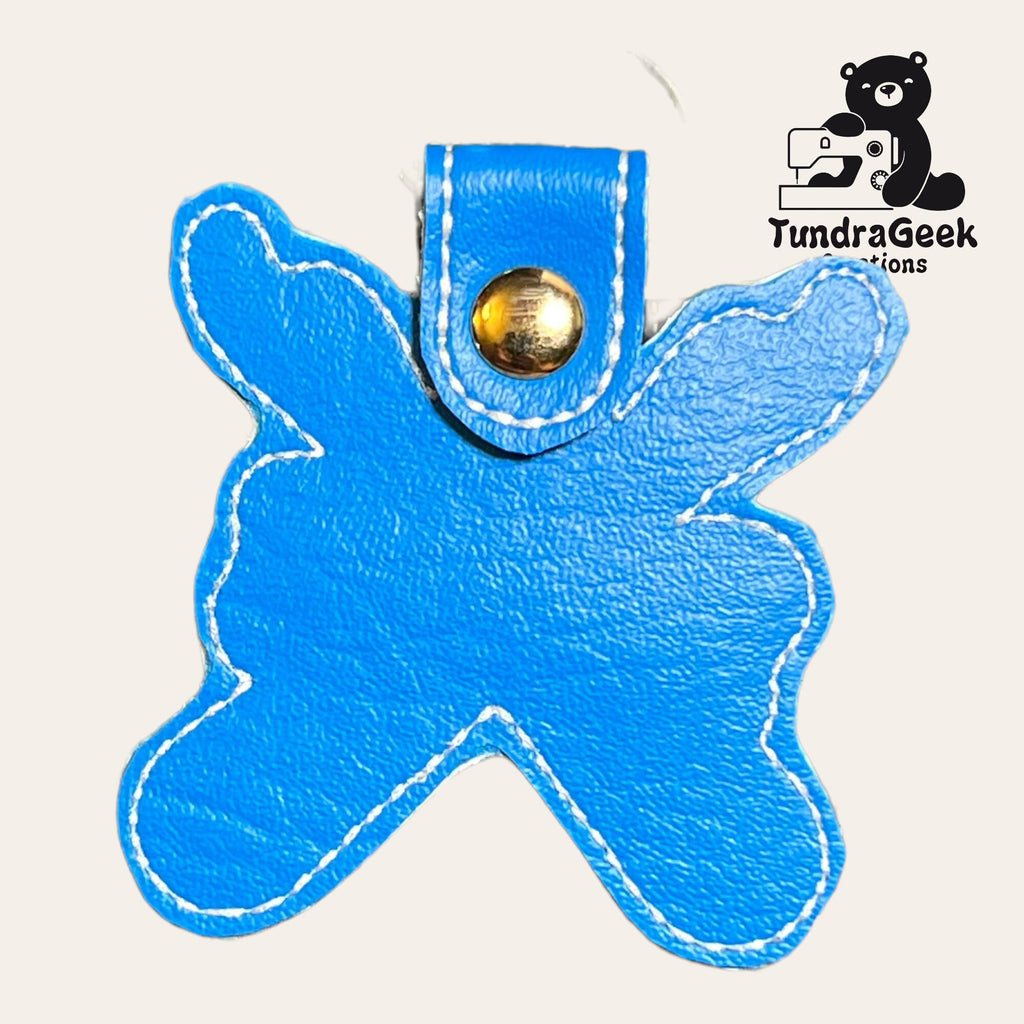 A blue, irregularly shaped leather keychain with white stitching and a gold snap button, shown next to the TundraGeek Creations logo featuring a sewing machine and bear.