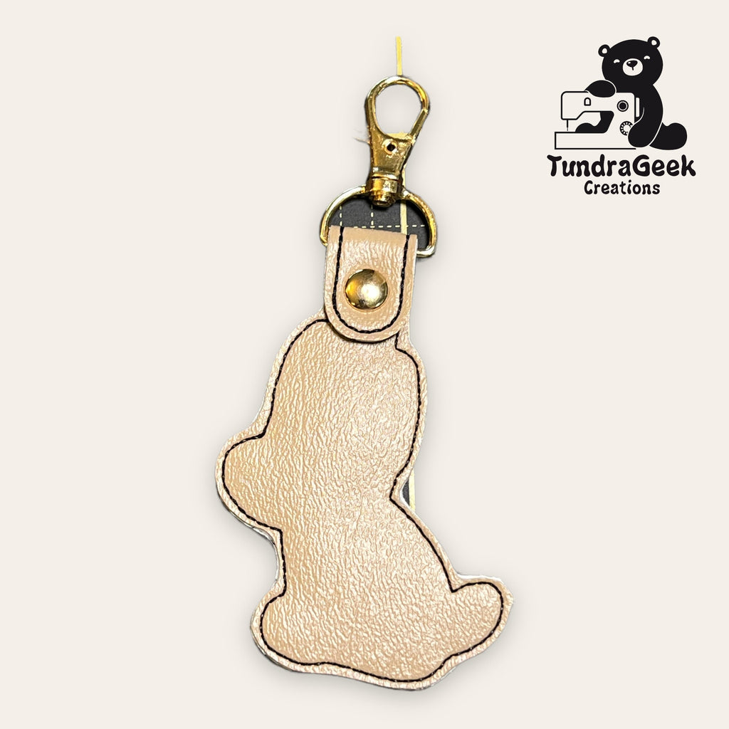 Tan leather keychain shaped like a bear, with a gold clasp at the top. A black bear and sewing machine logo with TundraGeek Creations appears in the top right corner.