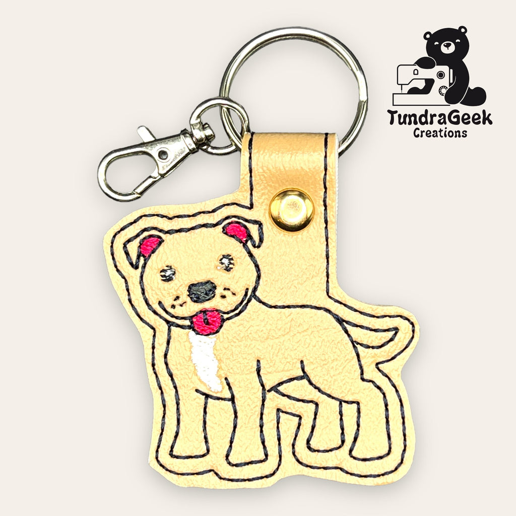 Tan faux leather keychain shaped like a cartoon dog with a pink collar, attached to a key ring and lobster clasp. TundraGeek Creations logo is in the top right corner.