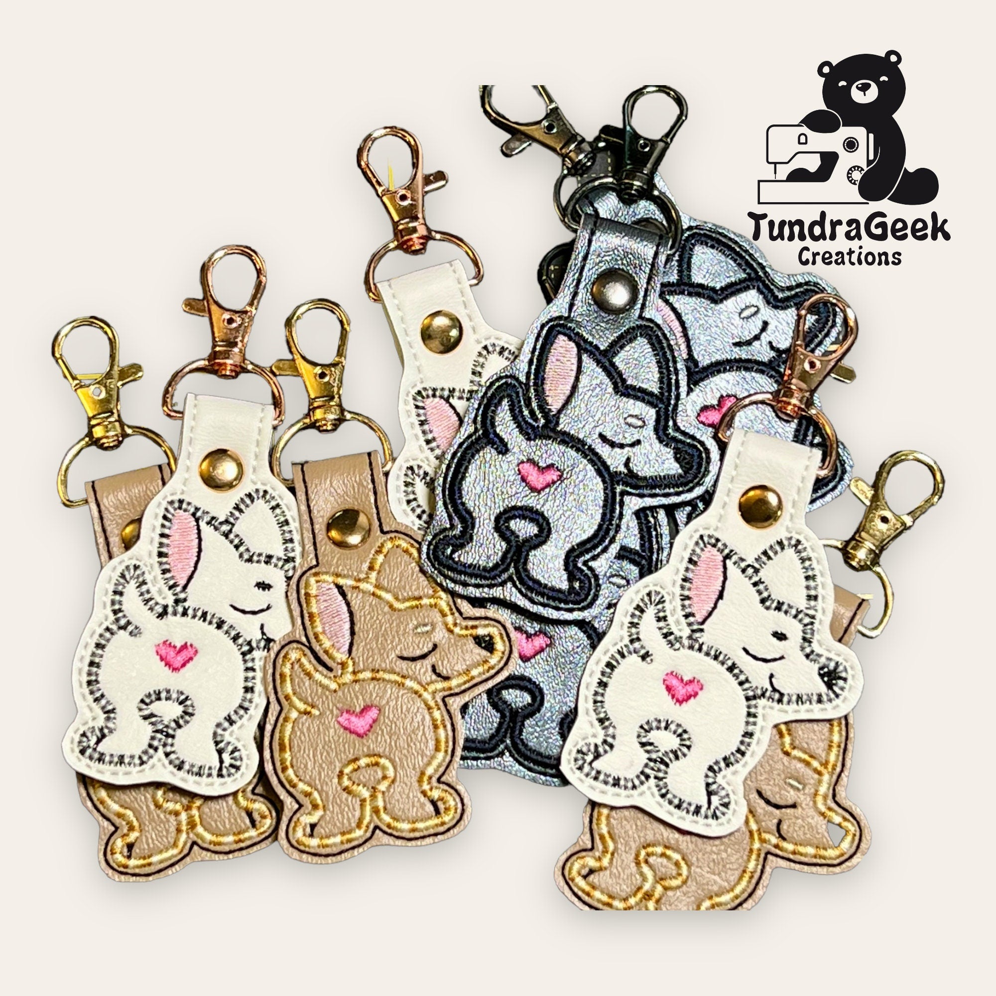 A set of embroidered keychains featuring cartoon bunnies with pink hearts, displayed in gold and silver hardware, next to a TundraGeek Creations logo with a bear and sewing machine.