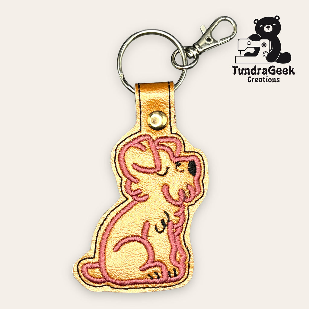 Dog-shaped embroidered keychain with a gold snap and metal clasp, featuring a pink outline and a TundraGeek Creations logo with a bear and sewing machine in the corner.
