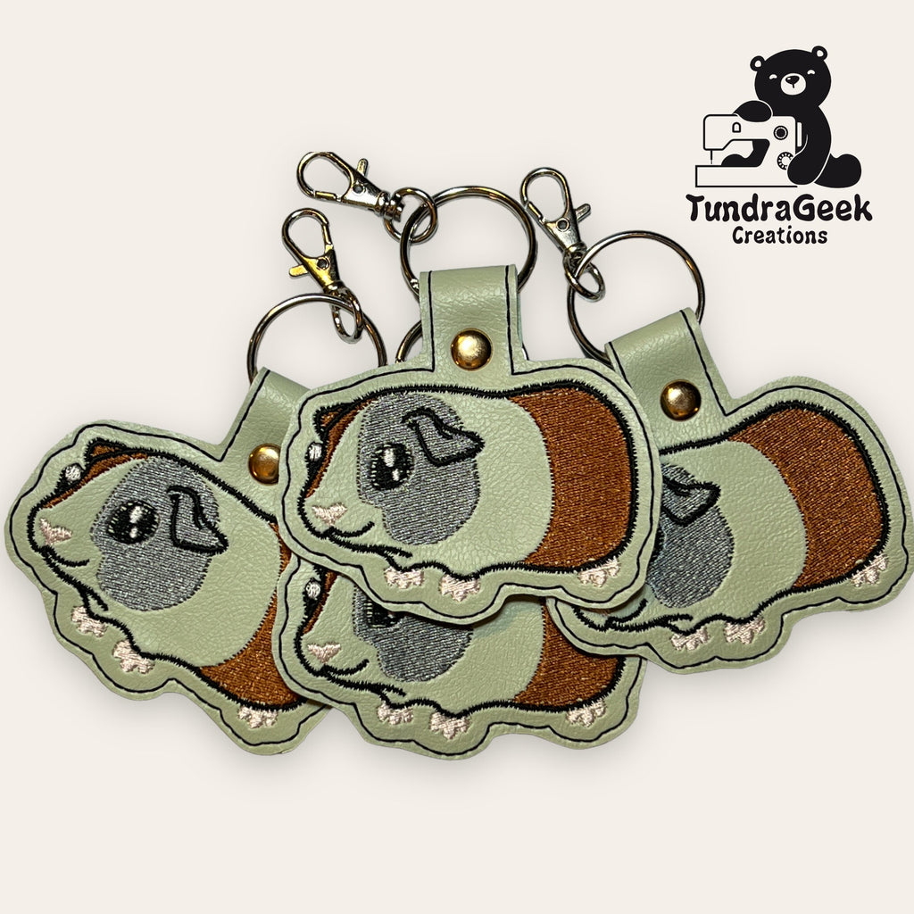 Four embroidered guinea pig keychains with metal clasps are arranged in a fan shape. The TundraGeek Creations logo appears in the top right corner.