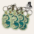 A group of embroidered keychains featuring teal and silver dragon designs, displayed with metal key rings and clasps. TundraGeek Creations logo is visible in the top right corner.