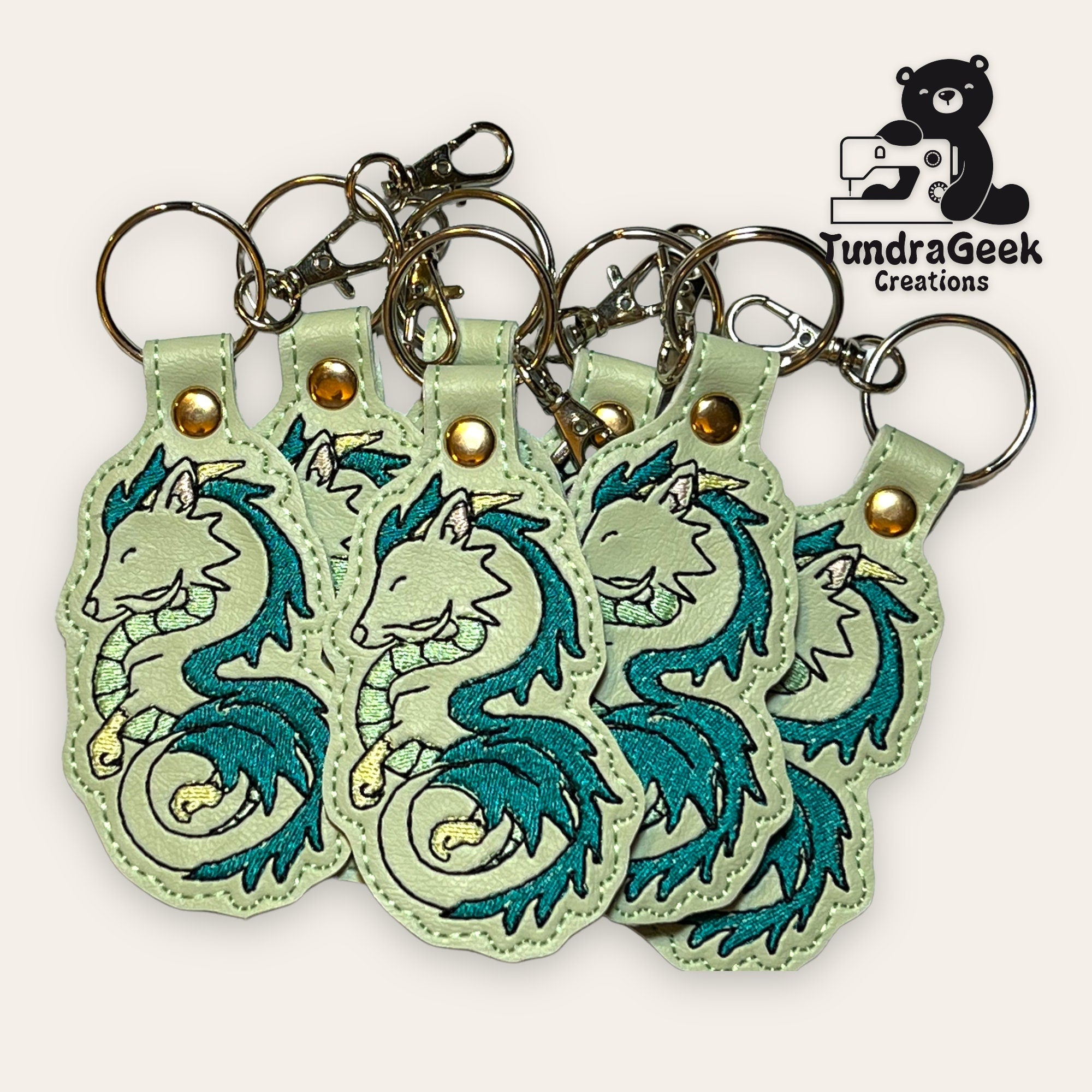 A group of embroidered keychains featuring teal and silver dragon designs, displayed with metal key rings and clasps. TundraGeek Creations logo is visible in the top right corner.