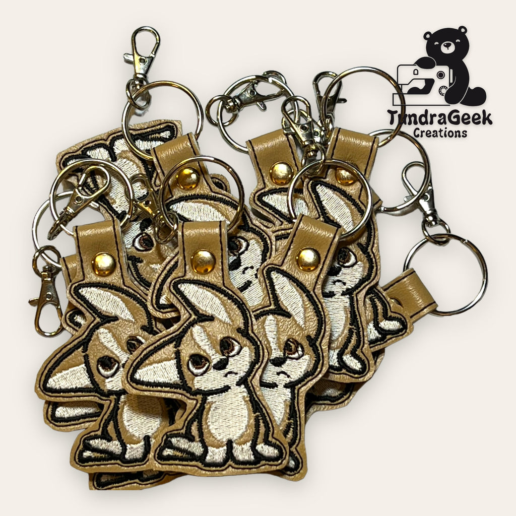 A cluster of embroidered keychains featuring a cartoon bunny design with metal rings and clasps, displayed on a light background. TundraGeek Creations logo is visible in the corner.