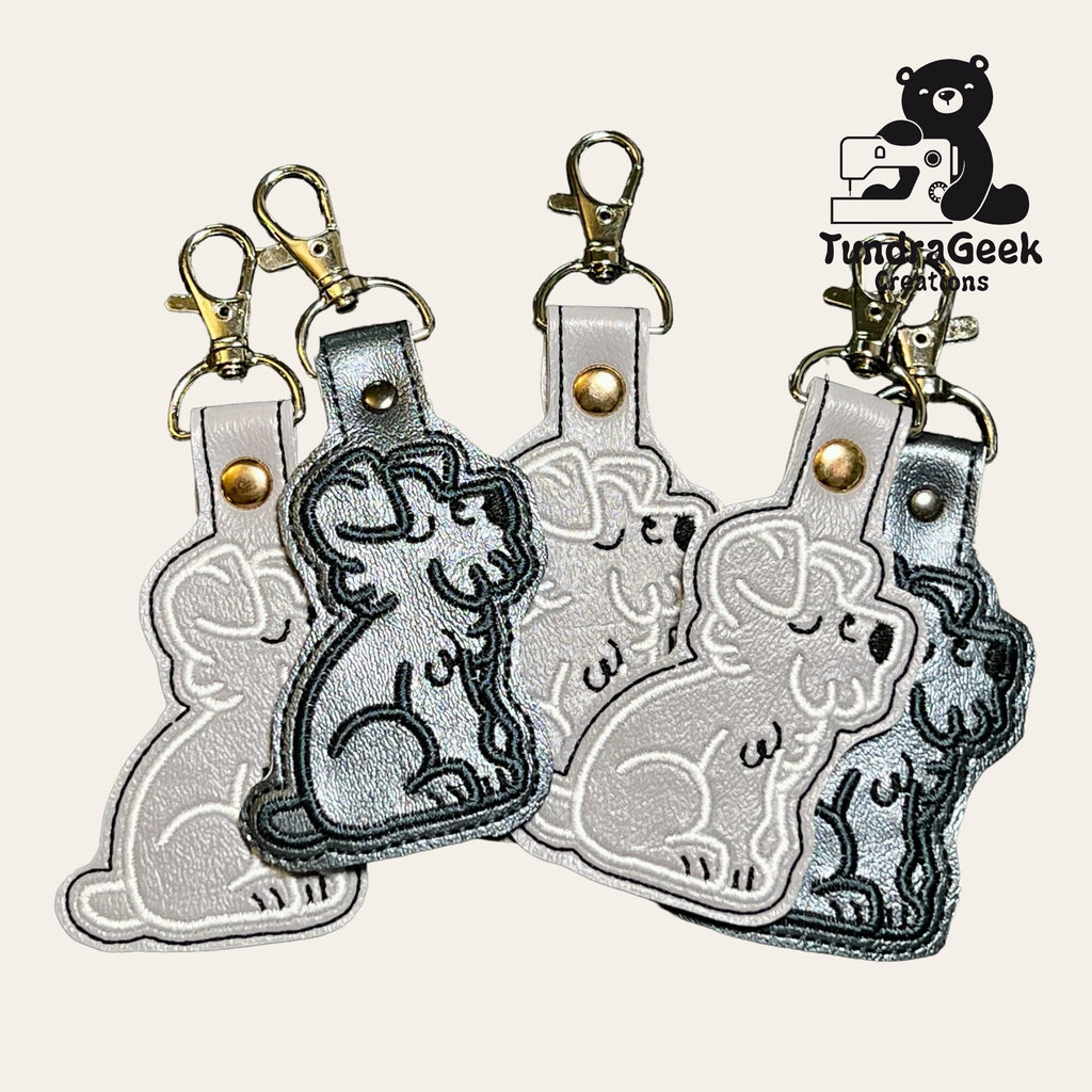 Four dog-shaped leather keychains with metal clasps are arranged in a fan pattern. A TundaGeek Creations logo featuring a bear with a sewing machine appears in the top right corner.