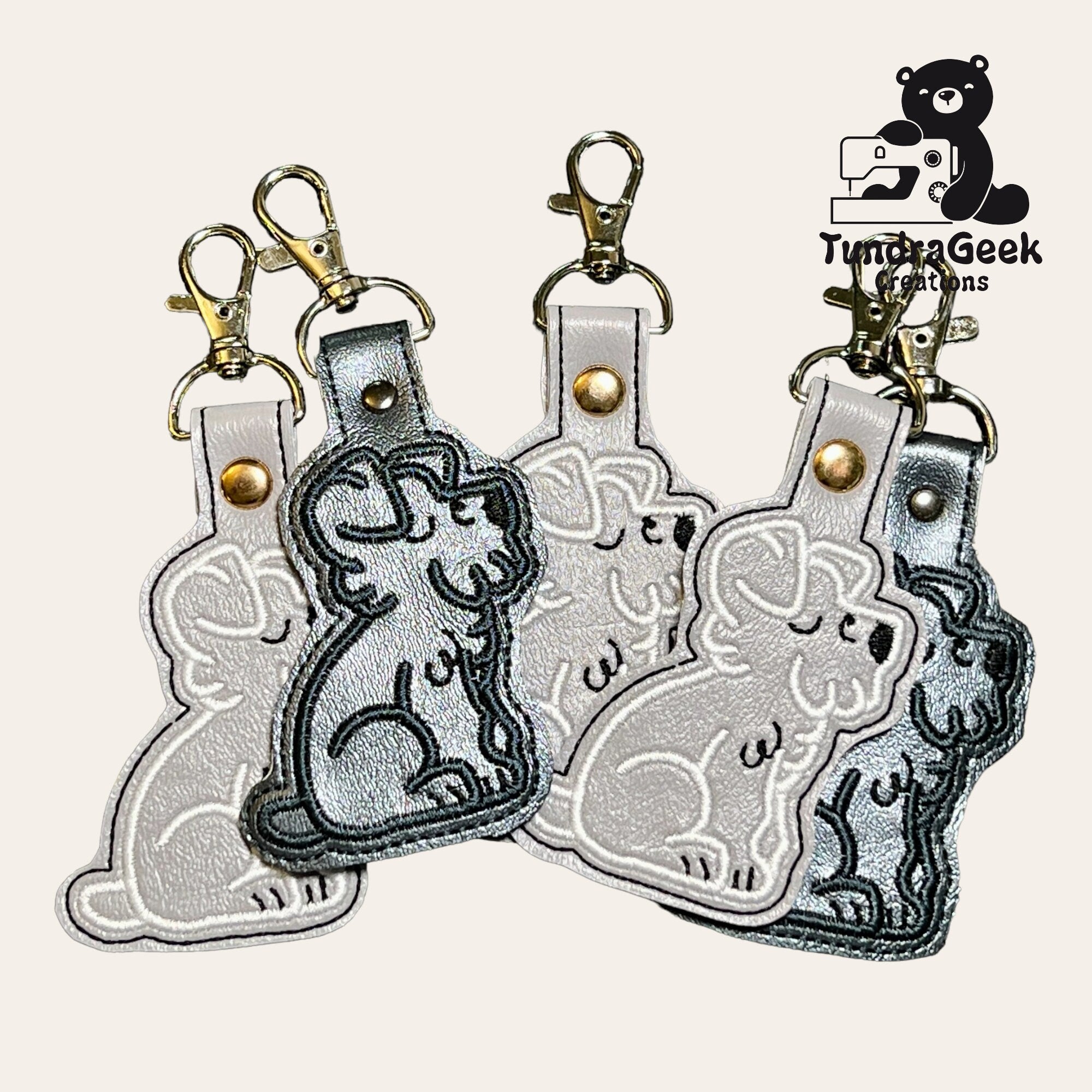 Four dog-shaped leather keychains with metal clasps are arranged in a fan pattern. A TundaGeek Creations logo featuring a bear with a sewing machine appears in the top right corner.