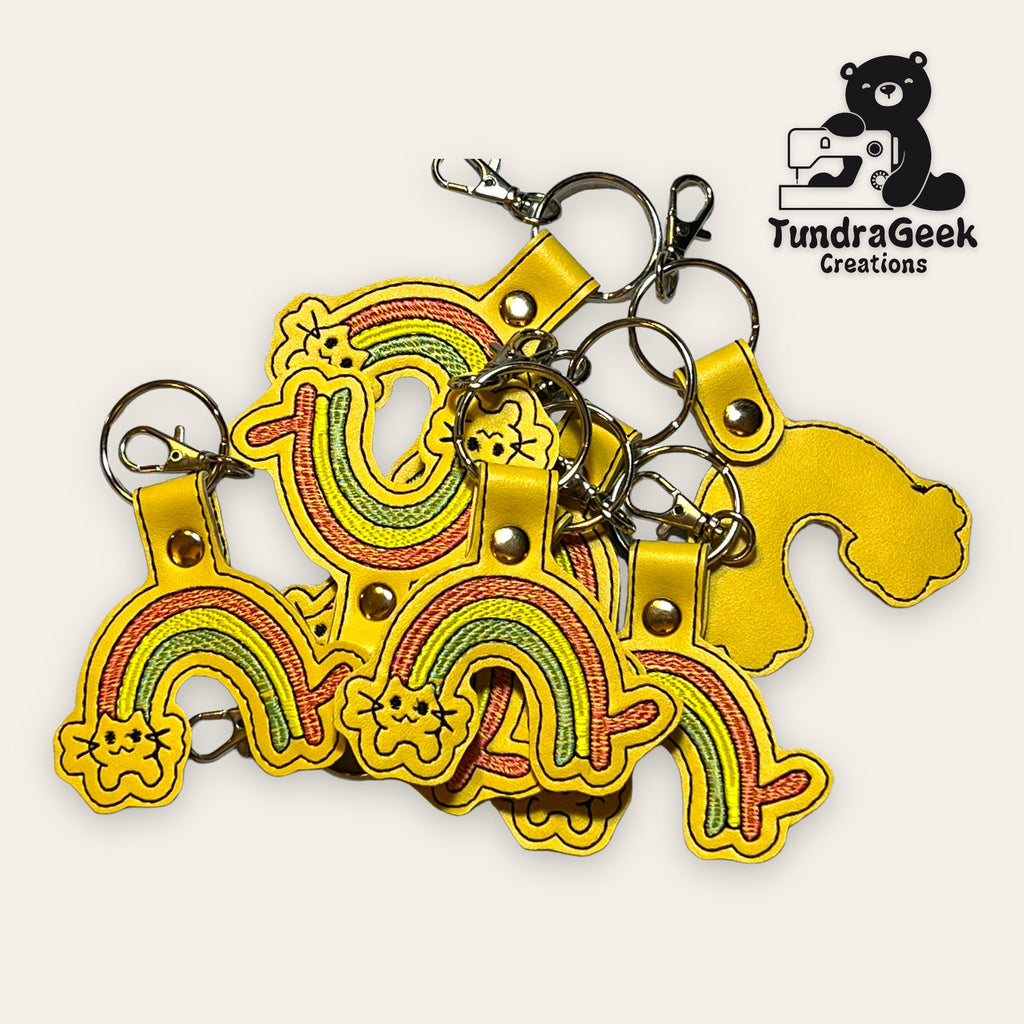 A pile of yellow rainbow-shaped keychains with embroidery, metal key rings, and clasps, next to the TundraGeek Creations logo featuring a bear with a sewing machine.