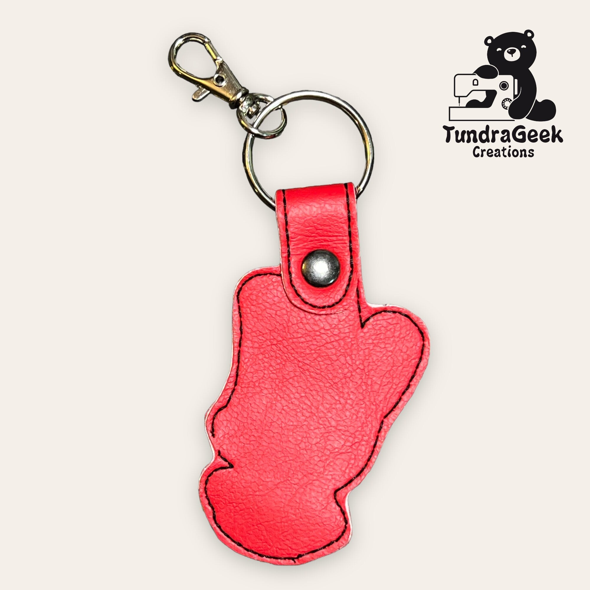 Red faux leather keychain in the shape of a mitten with a snap button and metal clasp, displayed on a light background with the TundraGeek Creations logo in the corner.